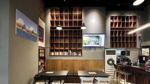 Ein gemütliches Café mit einer hölzernen Bank, Tischen und Stühlen. An der Wand befinden sich Regale und ein Fernseher.