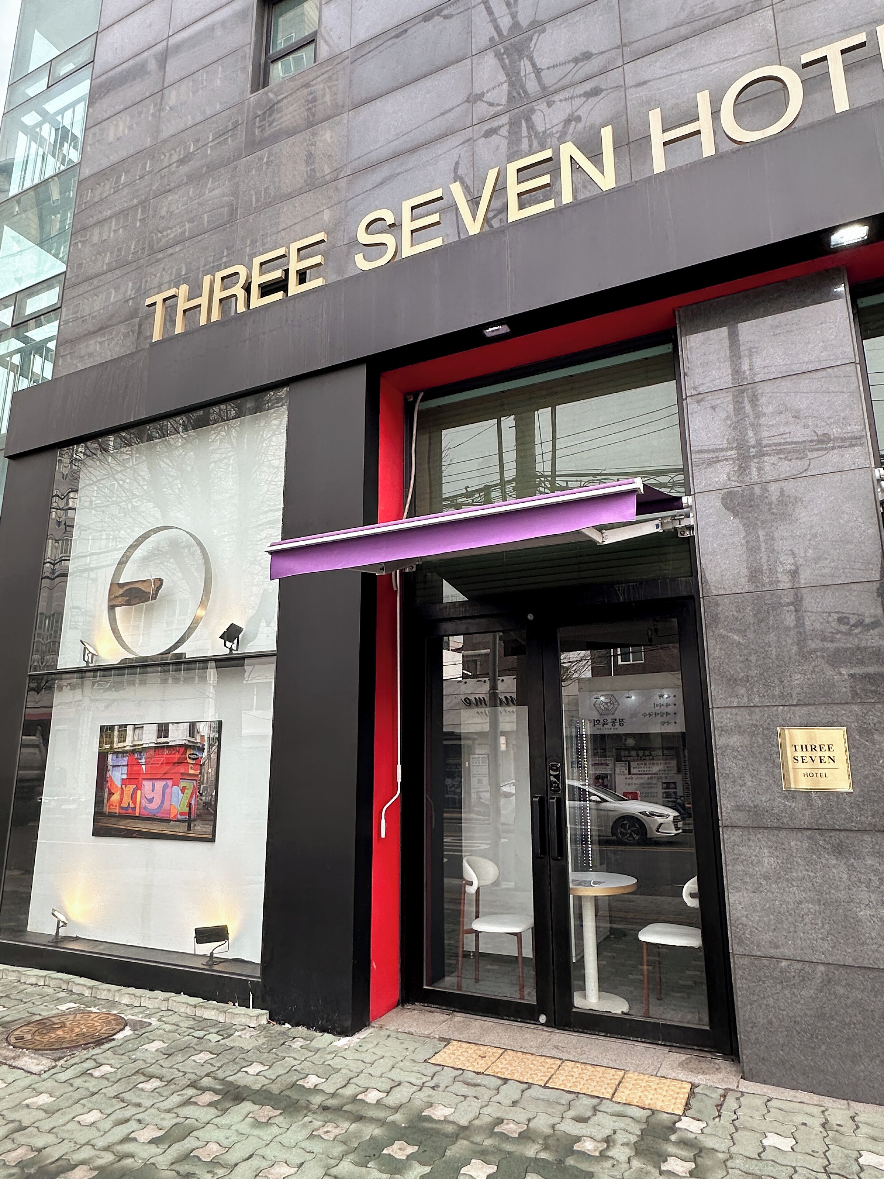 Fasaden på Three Seven Hotel är modern, med stora fönster och en tydligt markerad entré där det finns sittplatser.