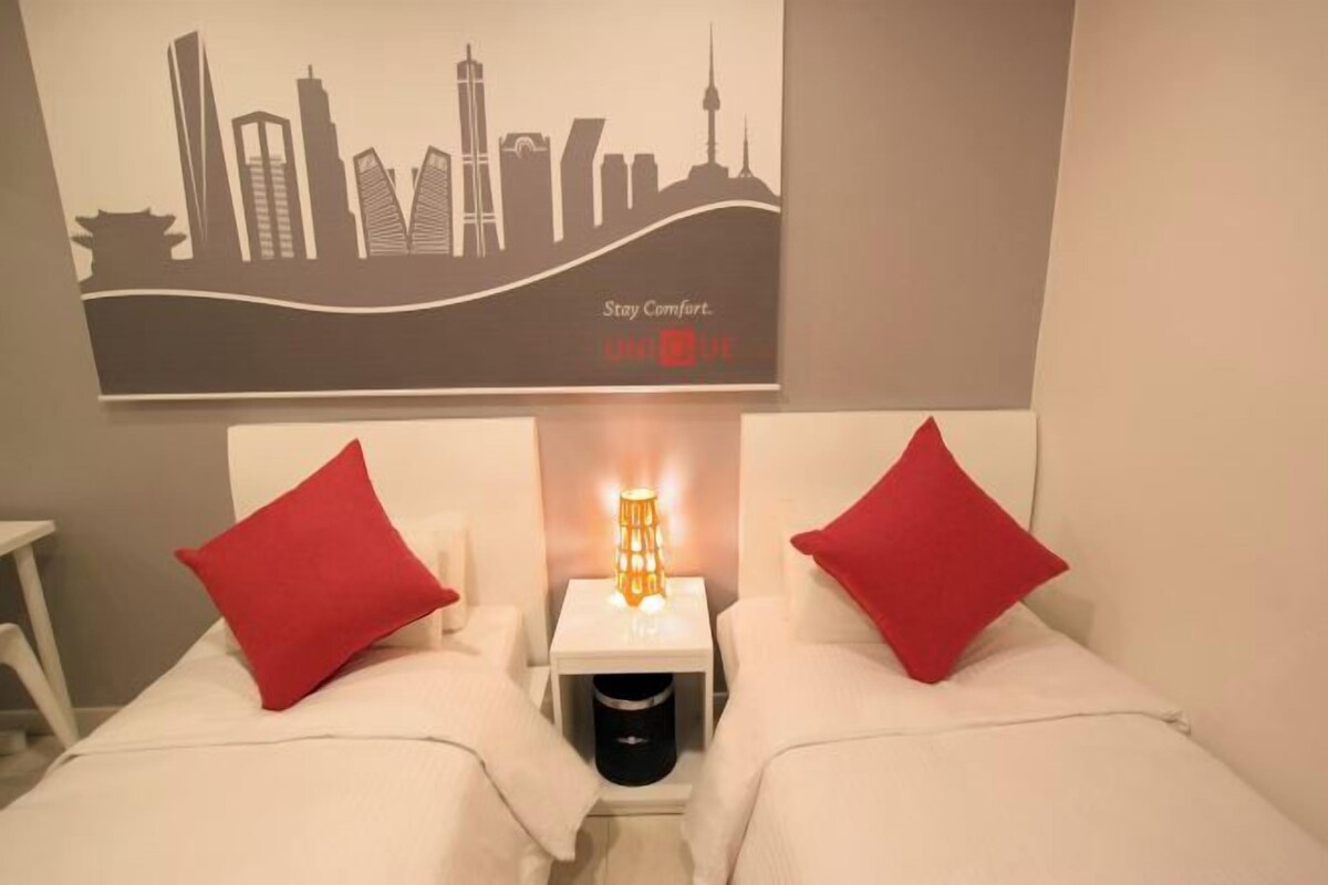 A hotel room with a bed, two red pillows, a nightstand with a lamp, and a city skyline poster on the wall.