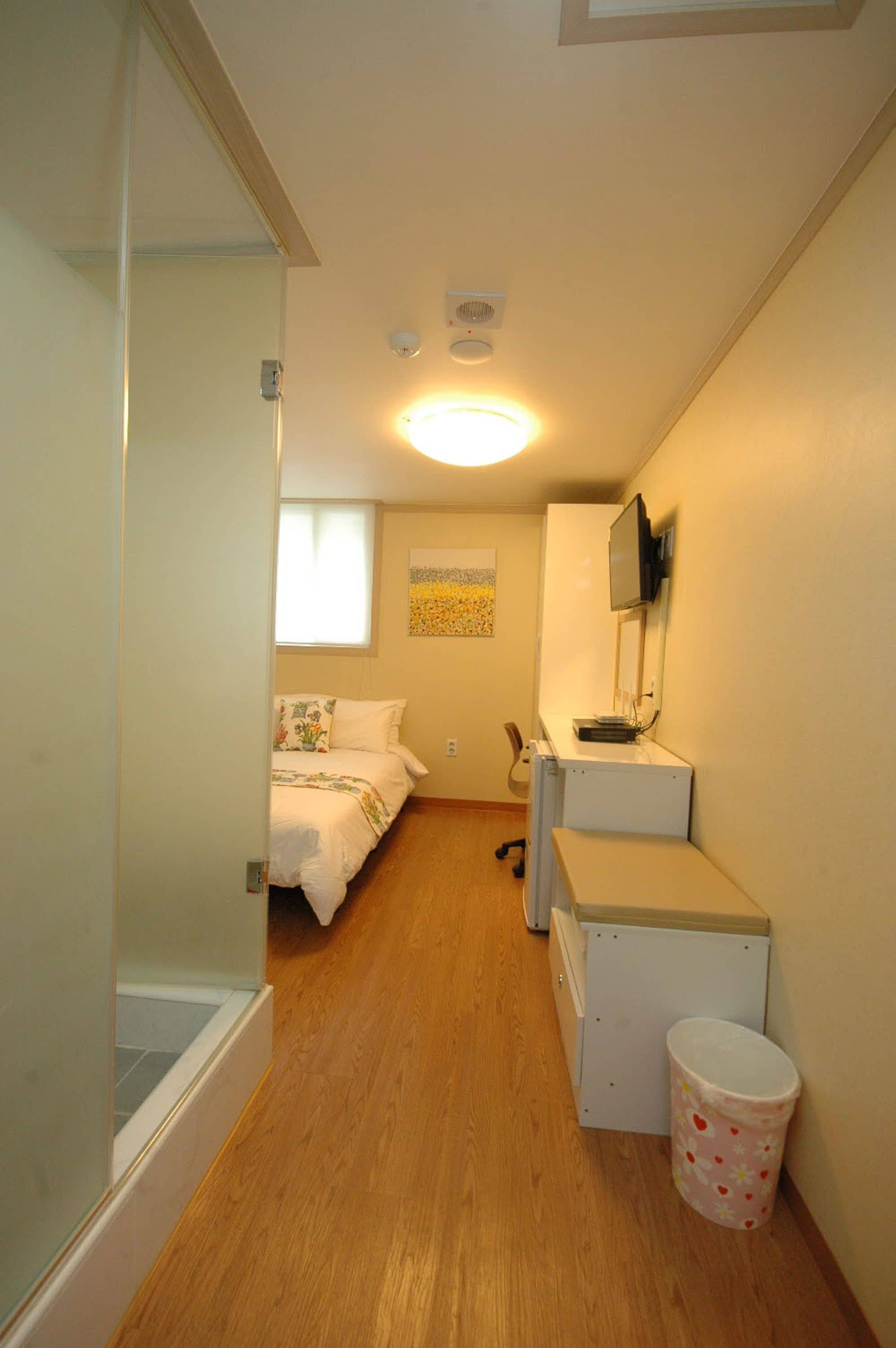 A narrow hallway with a bed, a desk, and a trash can.