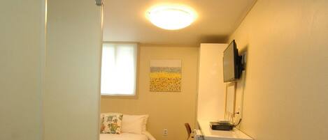 A narrow hallway with a bed, a desk, and a trash can.