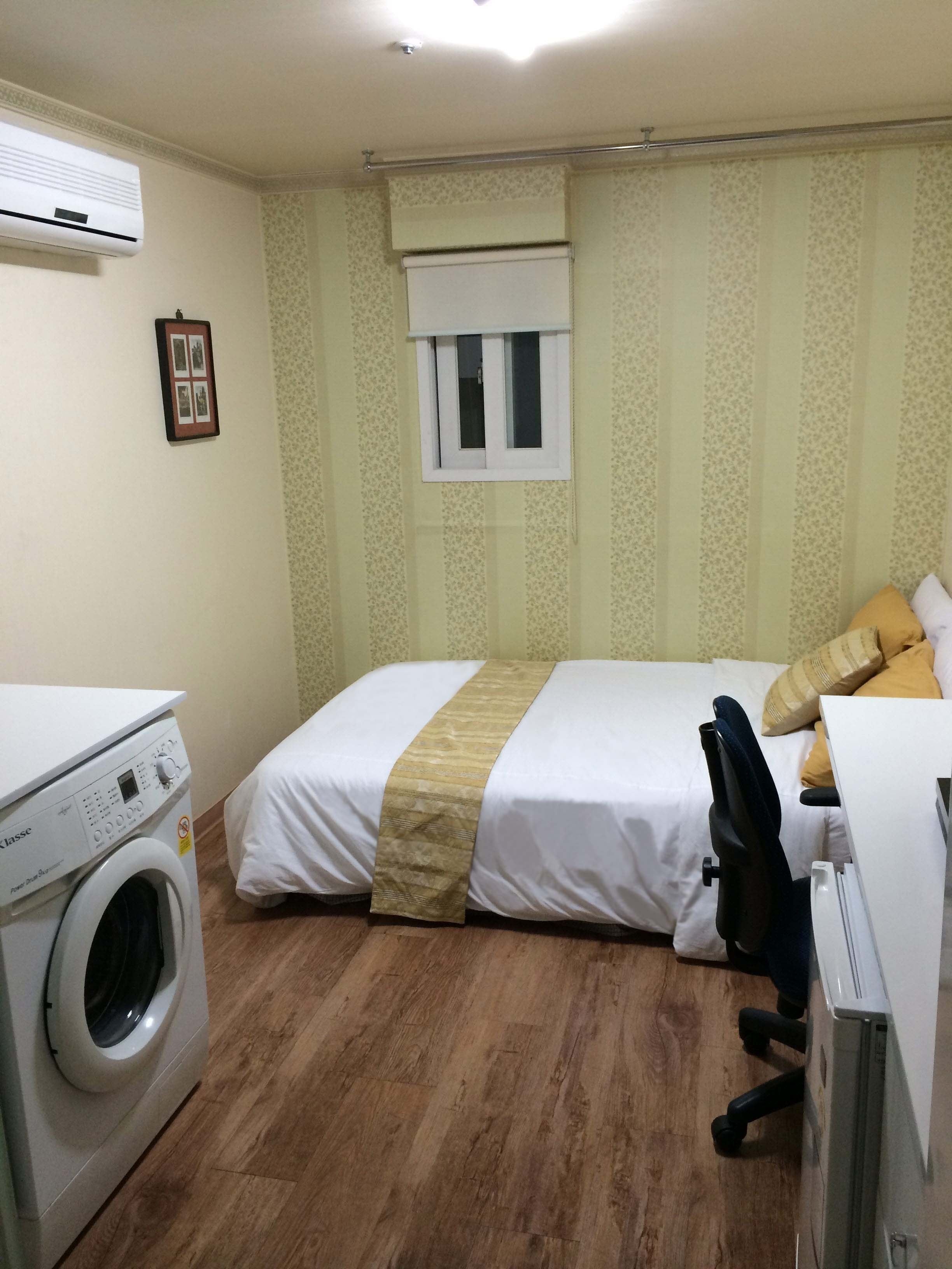 A hotel room with a bed, a desk, a chair, a washing machine, and a window with blinds.