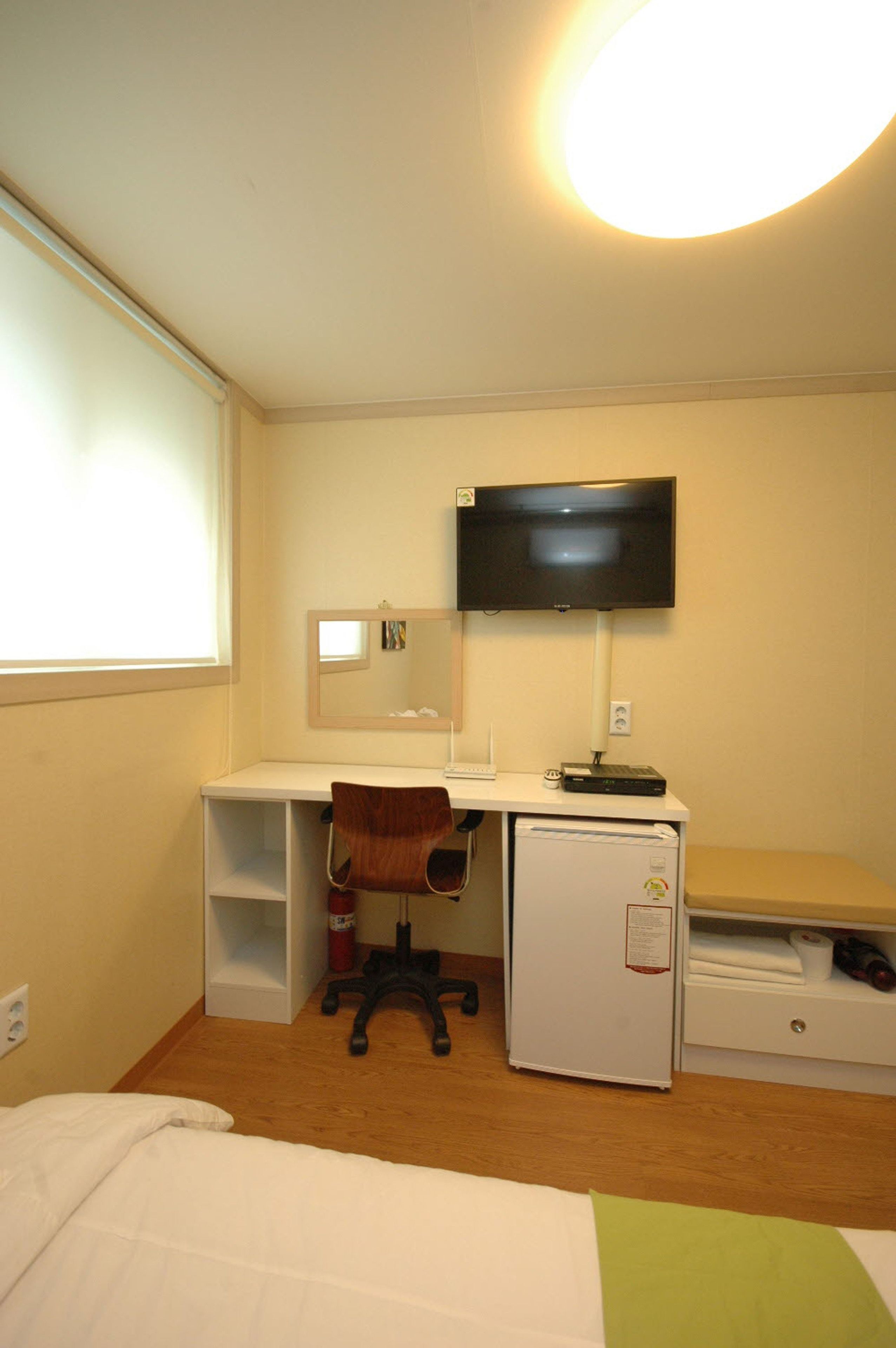 Una habitación de hotel con cama, escritorio y silla, televisor, refrigerador pequeño y ventana con persianas.