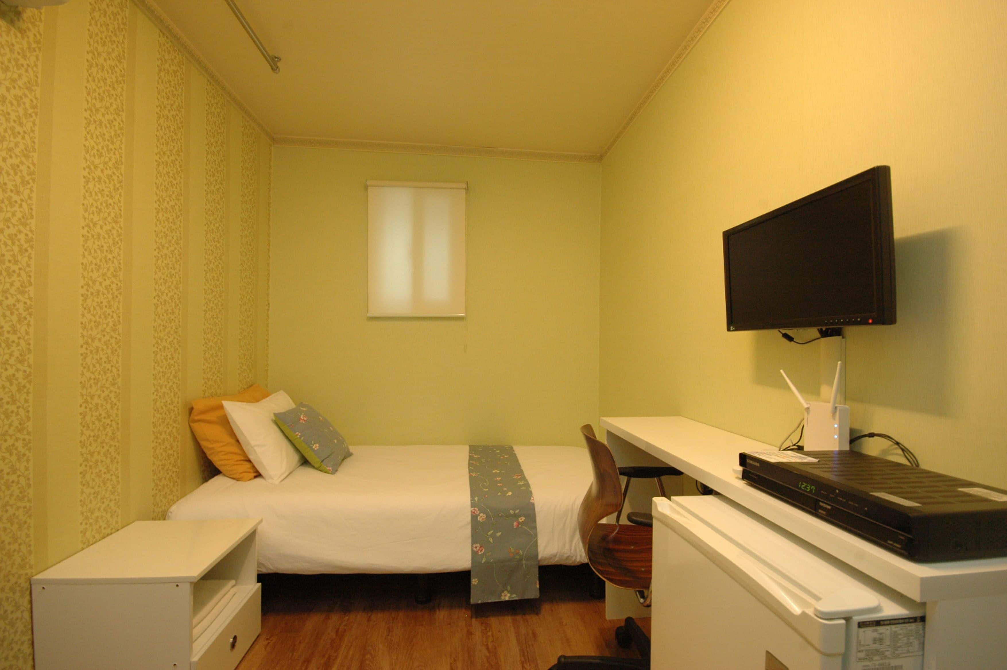 A small, neatly arranged bedroom with a single bed, a desk with a computer, and a television mounted on the wall.
