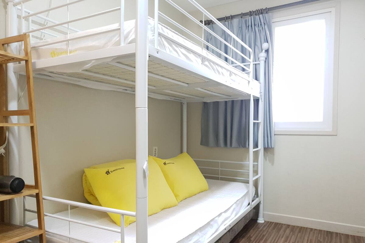 Habitación con literas, una escalera de madera, una ventana con cortinas y un estante con un objeto negro.