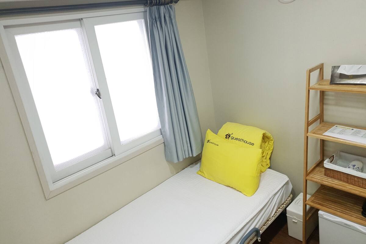 Una cama individual con colchón blanco y almohada amarilla con la etiqueta 'guesthouse'. Se ve una ventana con cortina azul, y a la derecha, un estante de madera con una cesta y un electrodoméstico blanco.