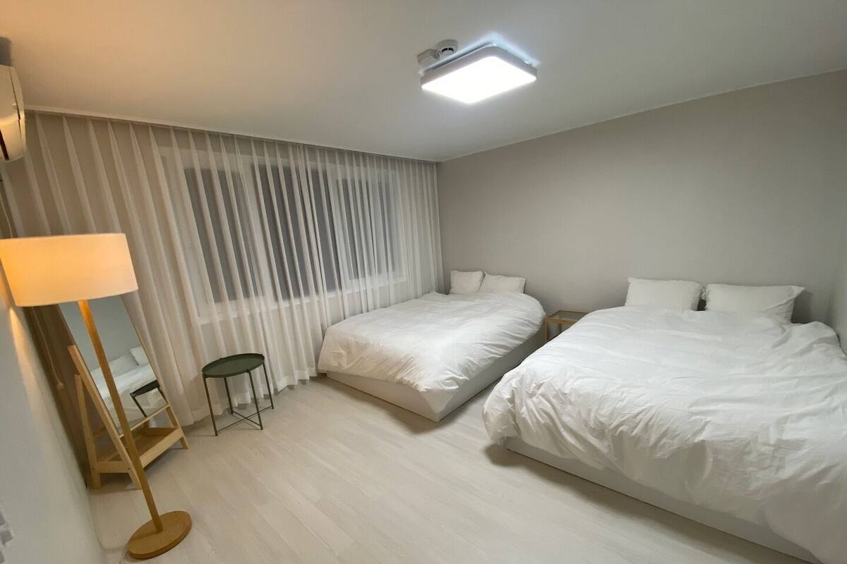 A hotel room with two beds, a standing lamp, a small table, and sheer curtains.