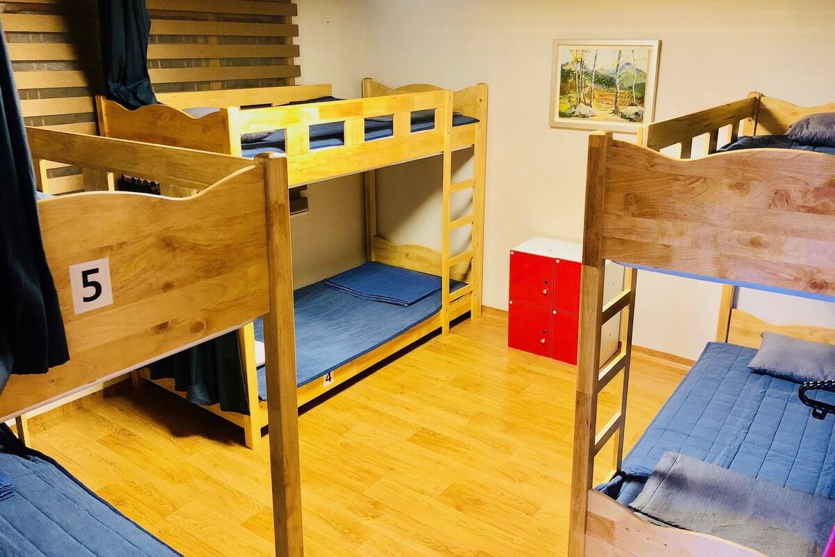 A dormitory room with bunk beds, wooden flooring, a red storage unit, and a painting on the wall.