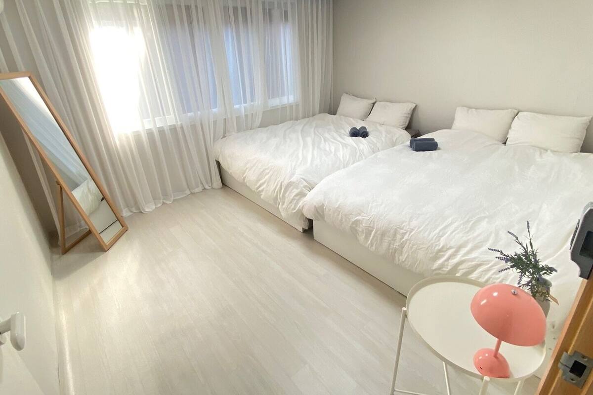 A bedroom with two beds, a mirror, a small table with a lamp, and a window with sheer curtains.