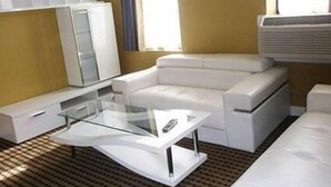 A modern hotel room with a white sofa, a glass coffee table, and a large window with blinds.