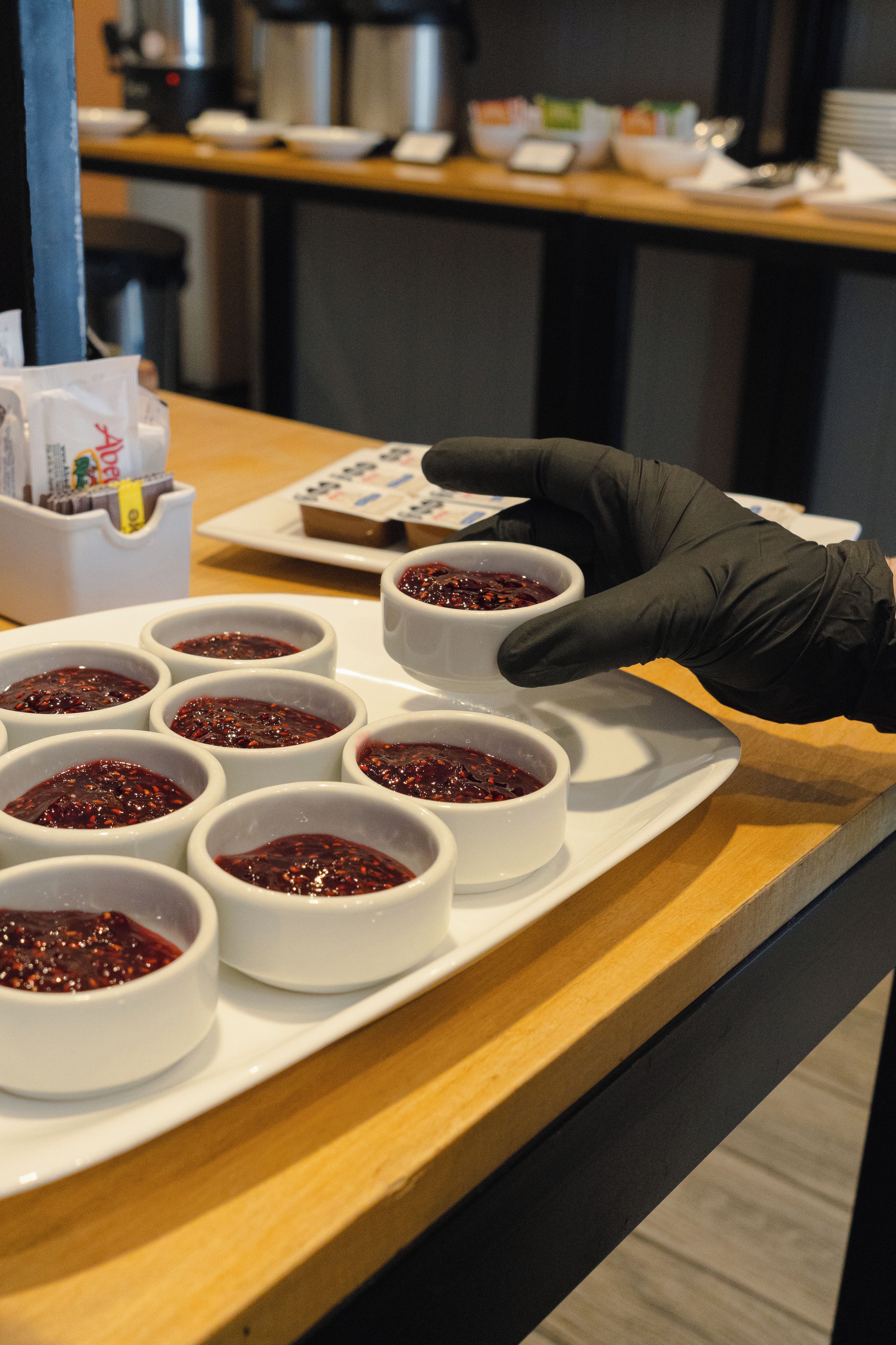 Una bandeja con varios recipientes de un condimento rojo oscuro, una persona con un guante negro manipulando los recipientes y una máquina de café al fondo.