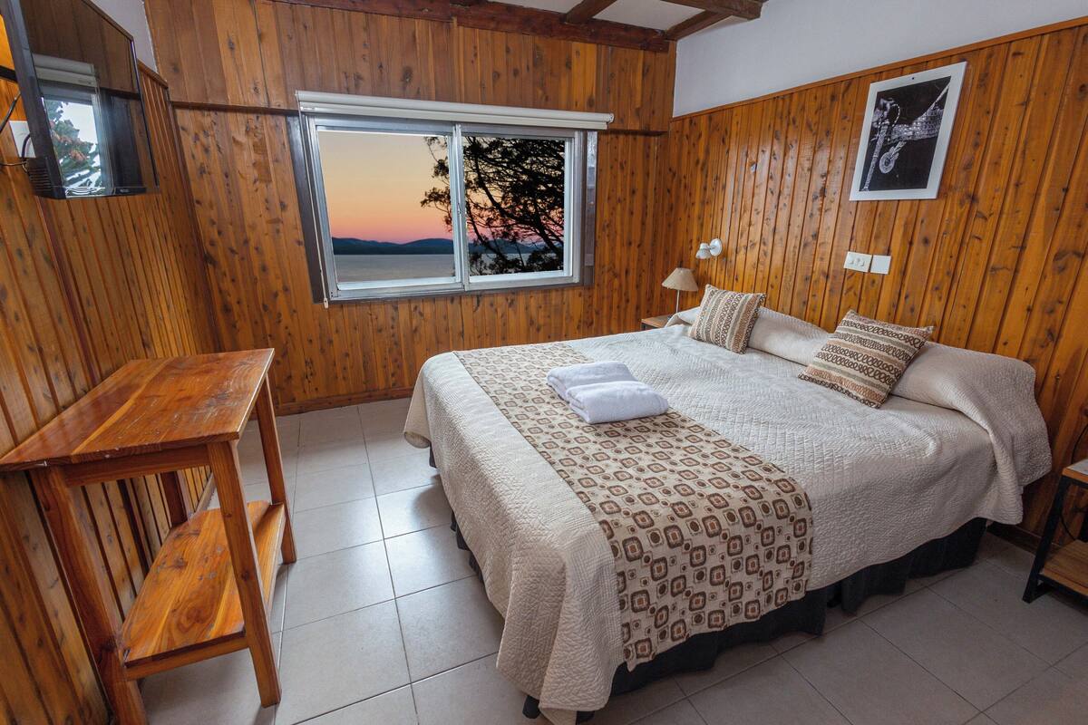 Una habitación con paneles de madera, una cama, un banco y un ventanal con vistas al atardecer.