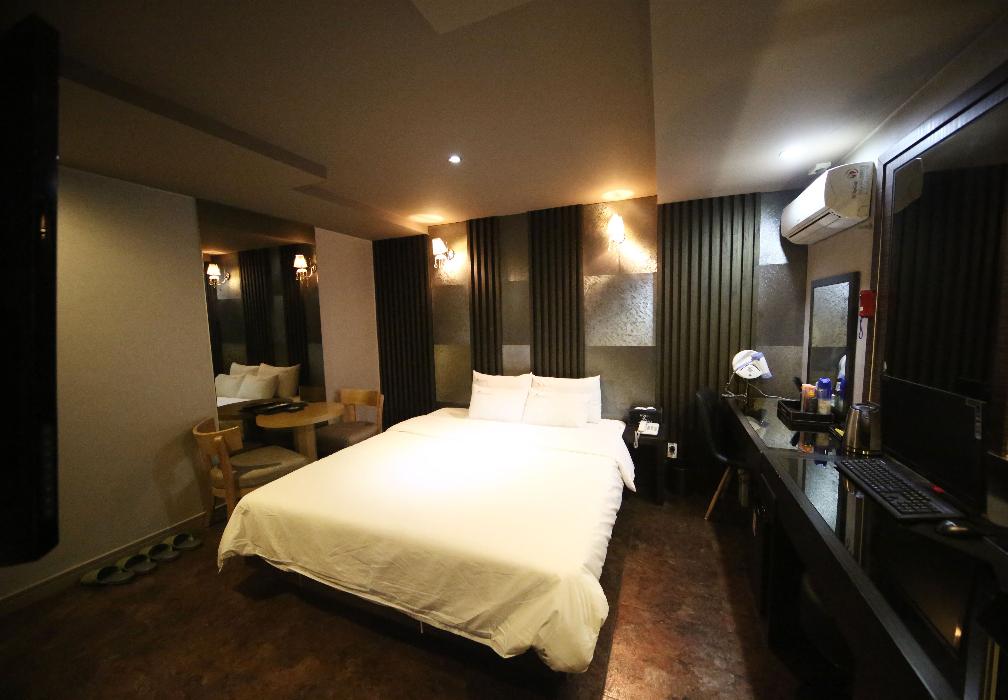 A hotel room with a large bed, a desk with a computer, a chair, a small table, and a mirror.