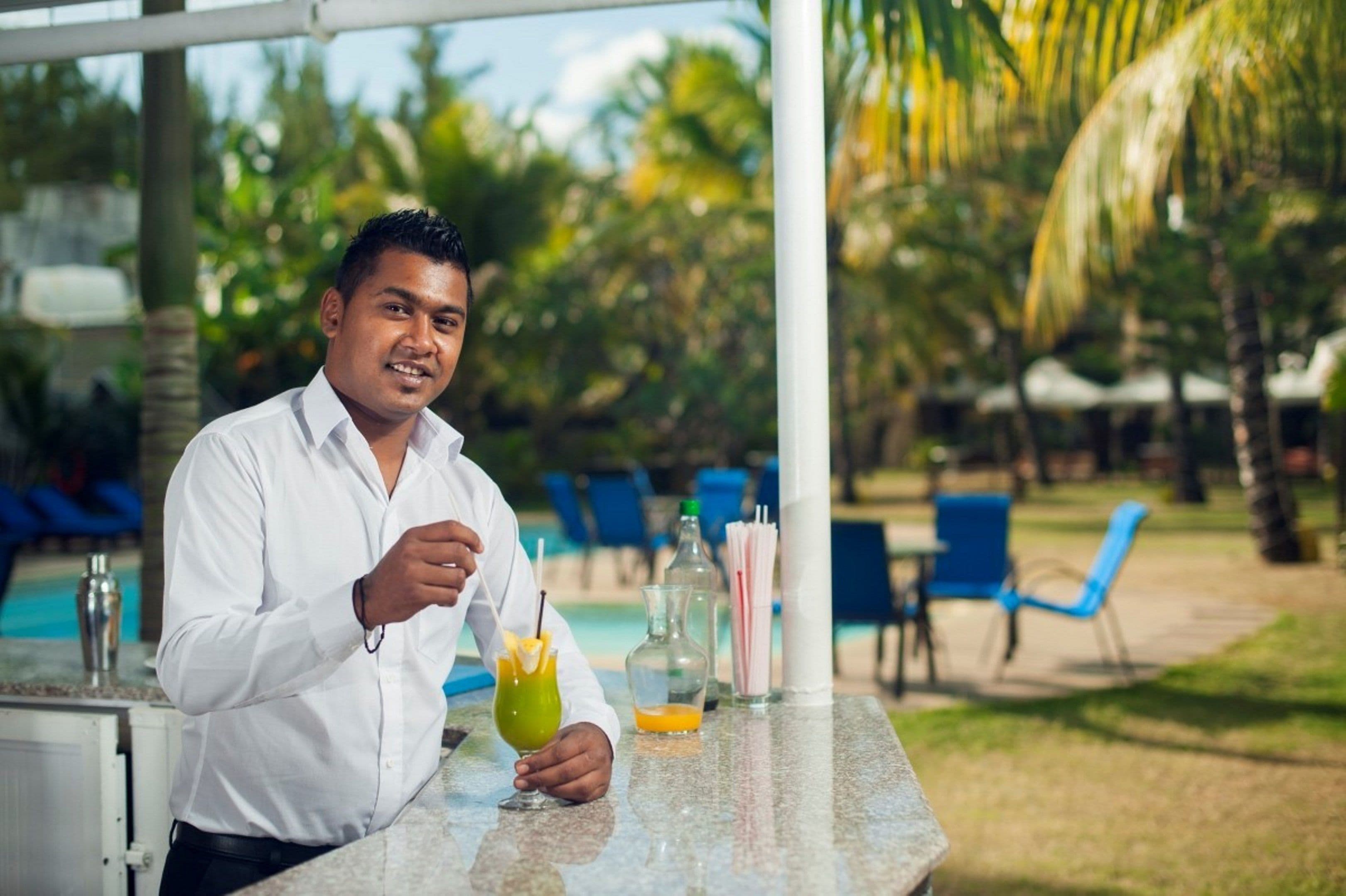 A person sitting at an outdoor bar with a drink, palm trees, and a pool in the background.
