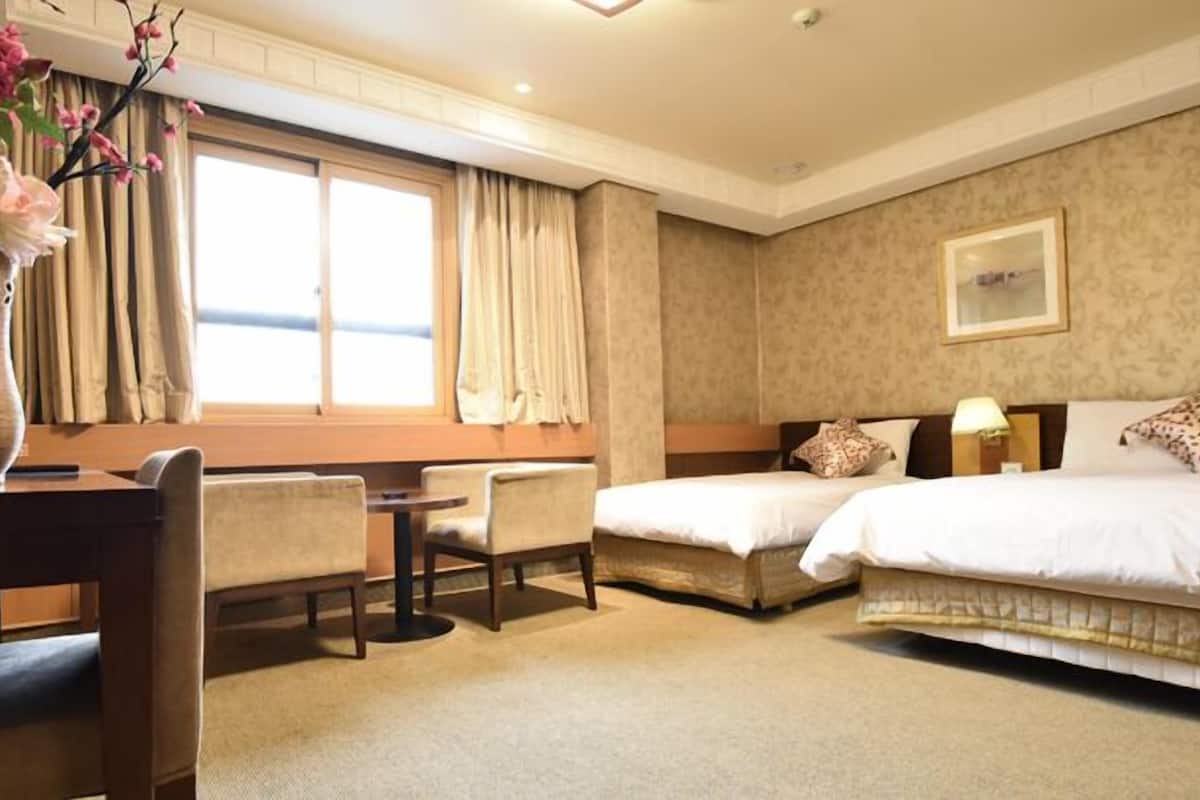 Habitación de hotel con dos camas, un escritorio con sillas, un florero con flores y una ventana con cortinas.