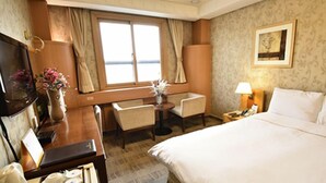 A hotel room with a bed, a desk, a chair, a small table, a lamp, and a window with curtains. - Hotel Samwon Plaza (Anyang)
