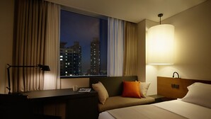 A hotel room with a bed, a desk, a chair, a lamp, and a view of city buildings at night. - Shilla Stay Dongtan (Hwaseong)
