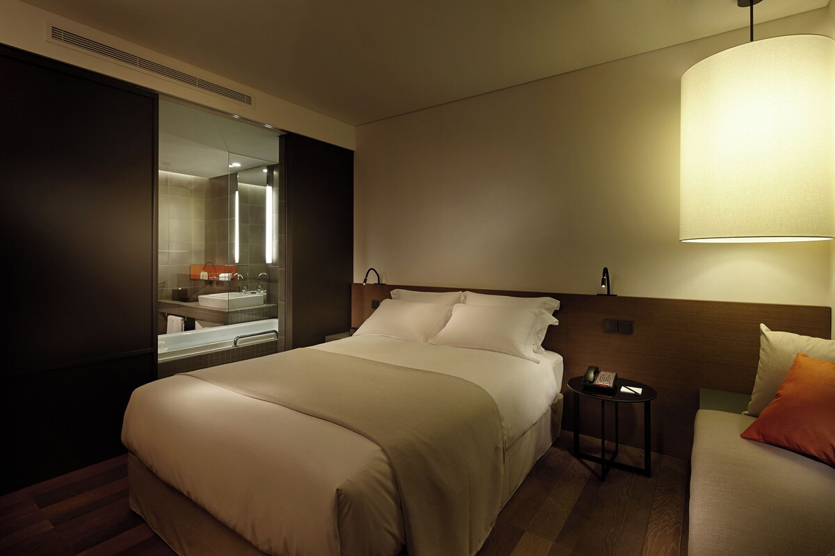 A hotel room with a large bed, a bedside table with a lamp, a mirror, and a view of the bathroom.