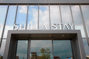 The exterior of a hotel named "SHILLA STAY" with large glass windows reflecting the sky and surrounding buildings. - Shilla Stay Dongtan (Hwaseong)