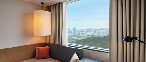 A hotel room with a bed, a desk, a lamp, and a view of a cityscape through a large window.