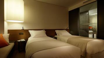A hotel room with two beds, a nightstand, a lamp, and a bathroom visible through a glass door.