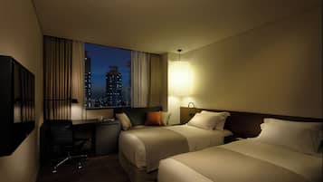 A hotel room with two beds, a desk, a chair, a lamp, and a view of a cityscape at dusk.