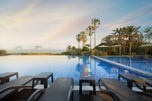 Zona junto a la piscina con sillones de descanso y una mesa, con vistas a una piscina cristalina y a islas distantes.