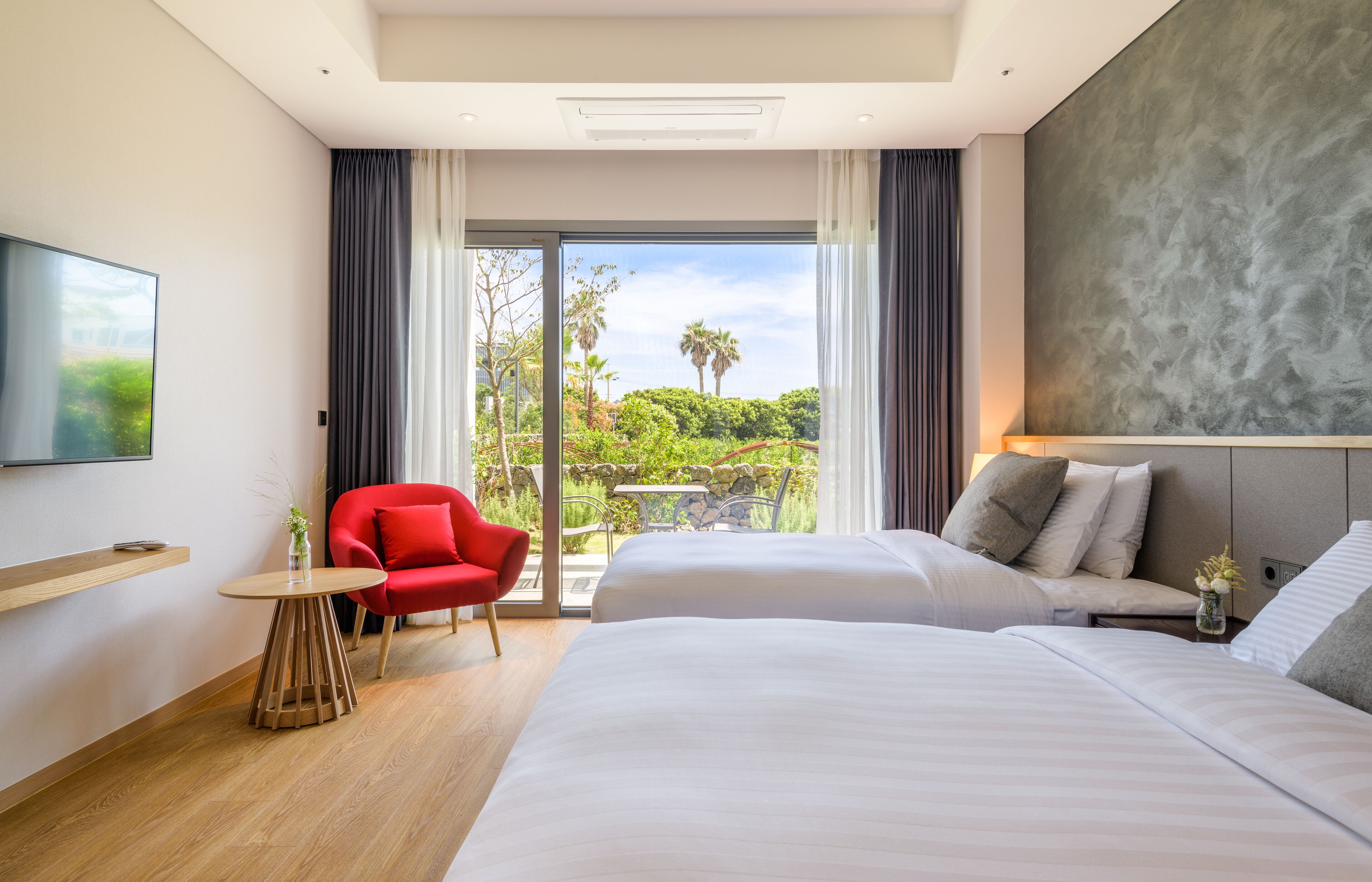 A hotel room with two beds, a red chair, a small table, and a large window offering a view of greenery.