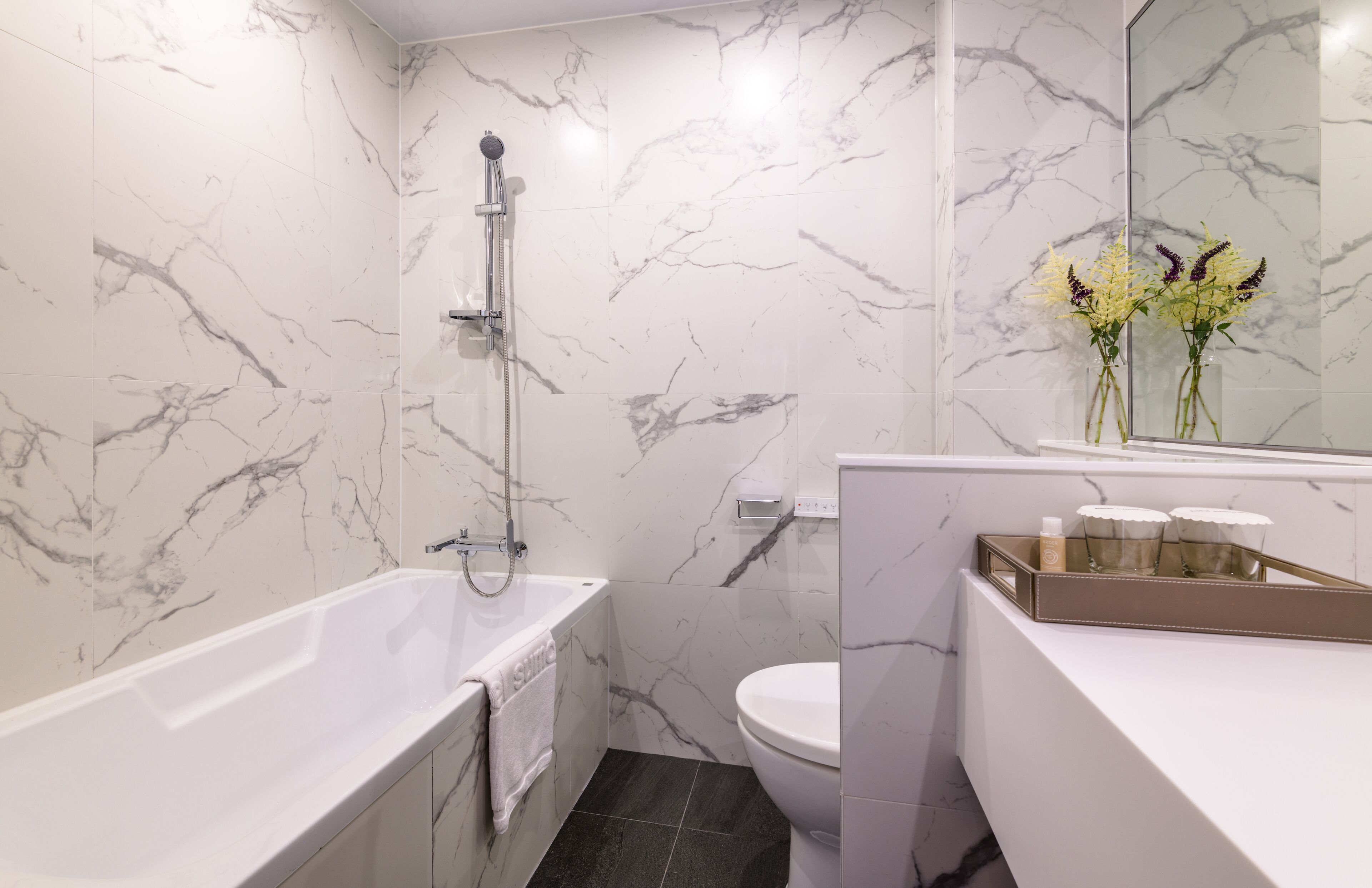 A modern bathroom with a marble wall, a bathtub, a toilet, and a vanity with a tray of toiletries.