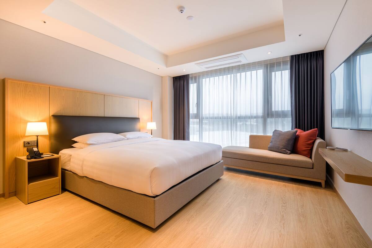 A modern hotel room with a large bed, bedside lamps, a sofa, and a large window with curtains.