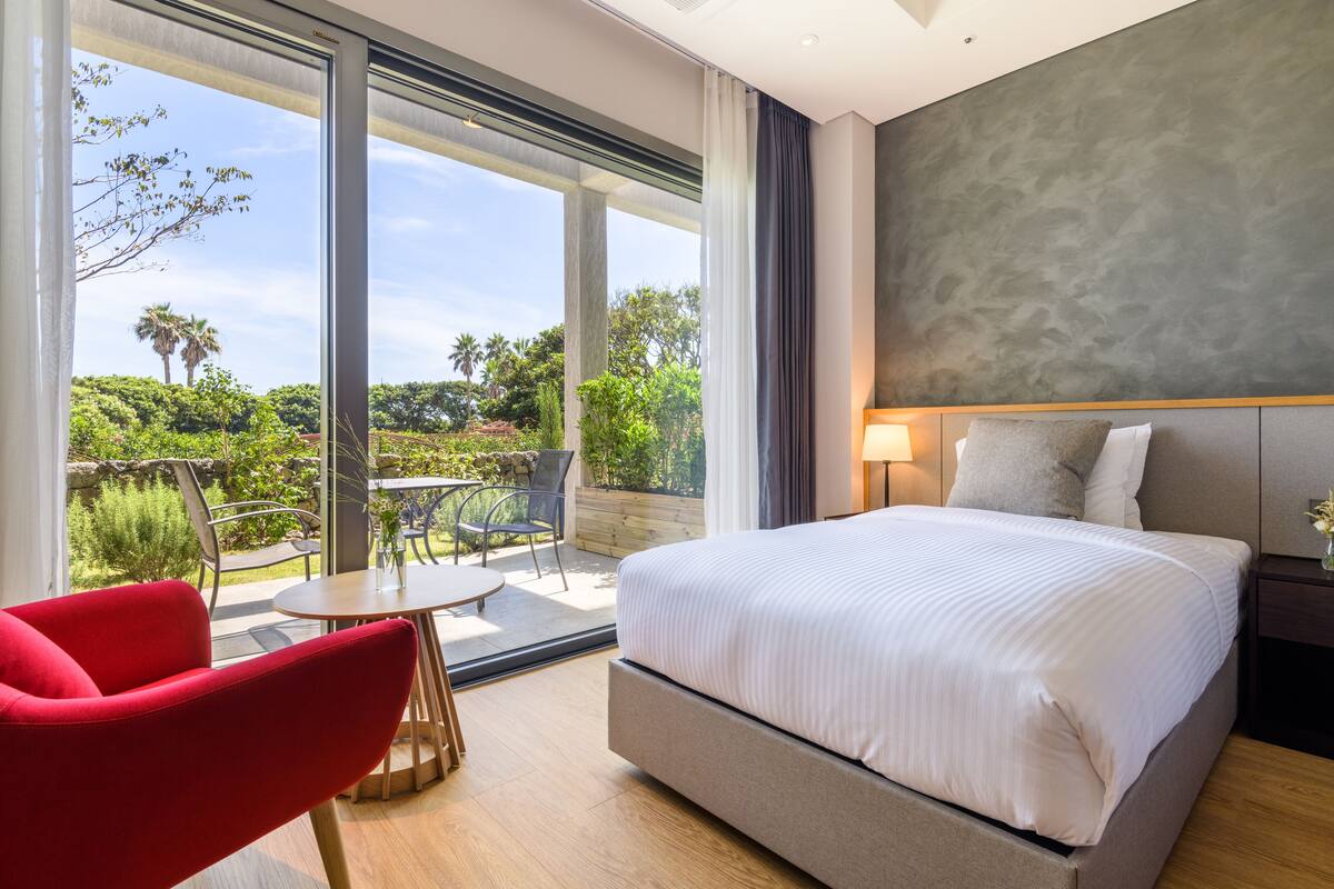 A modern hotel room with a large bed, a red chair, a small table, and a balcony with a view of greenery and palm trees.
