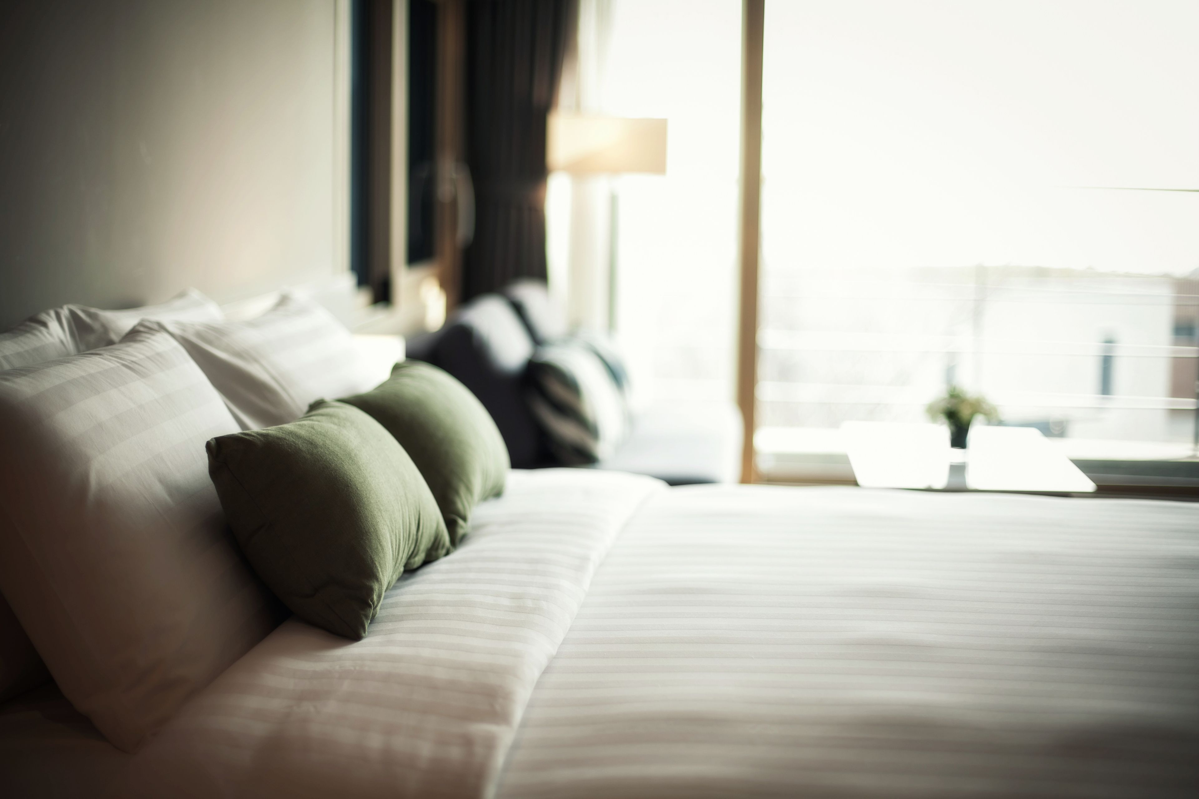 A neatly made bed with pillows, a bedside table with a lamp, and a window with a view of buildings.