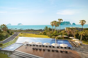 Una piscina infinita en la azotea con sillones de descanso y sombrillas, con vistas a una playa con palmeras y cielo despejado.