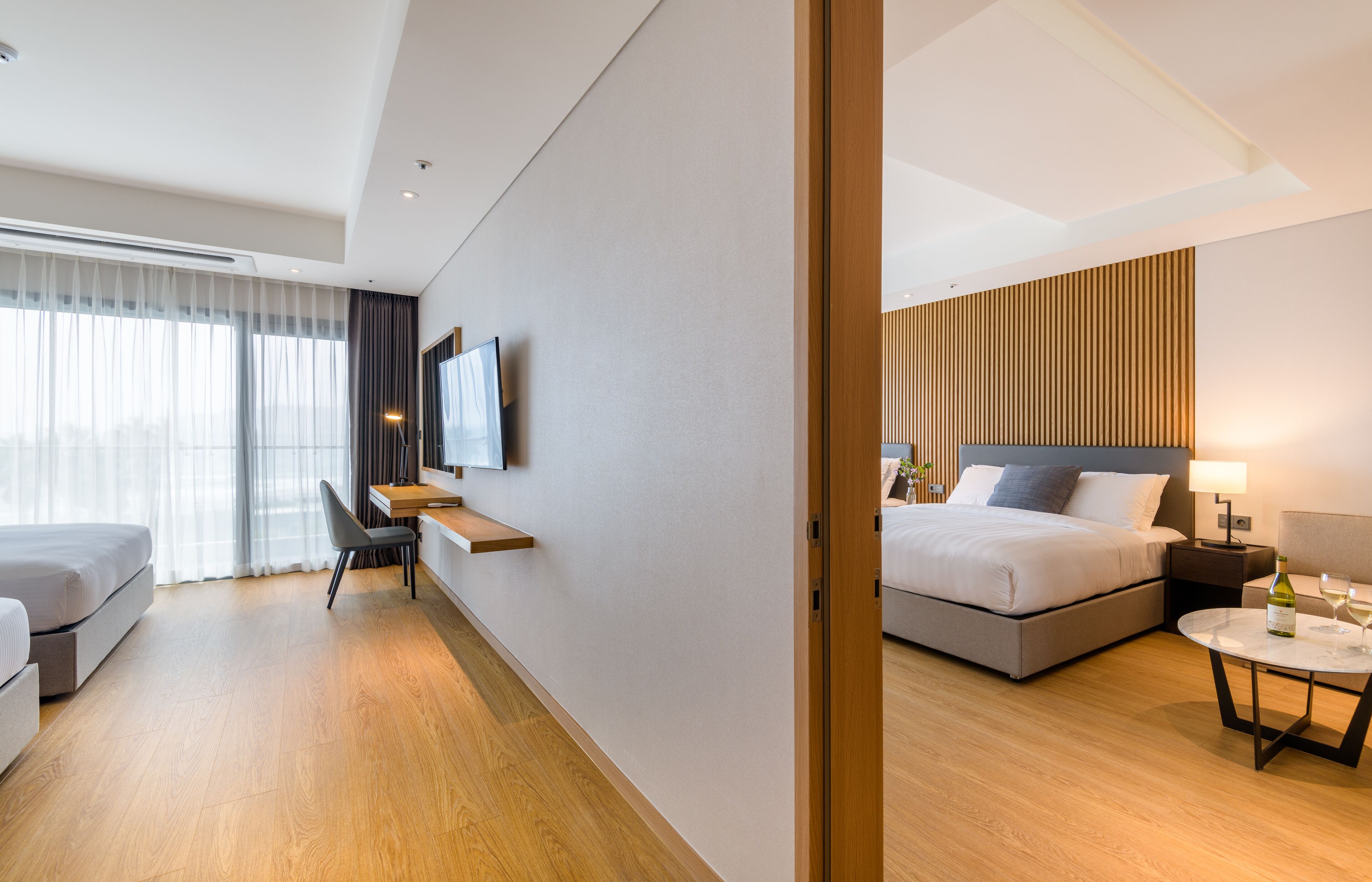 A modern hotel room with a bed, bedside table, lamp, and a small table with a bottle and glass.