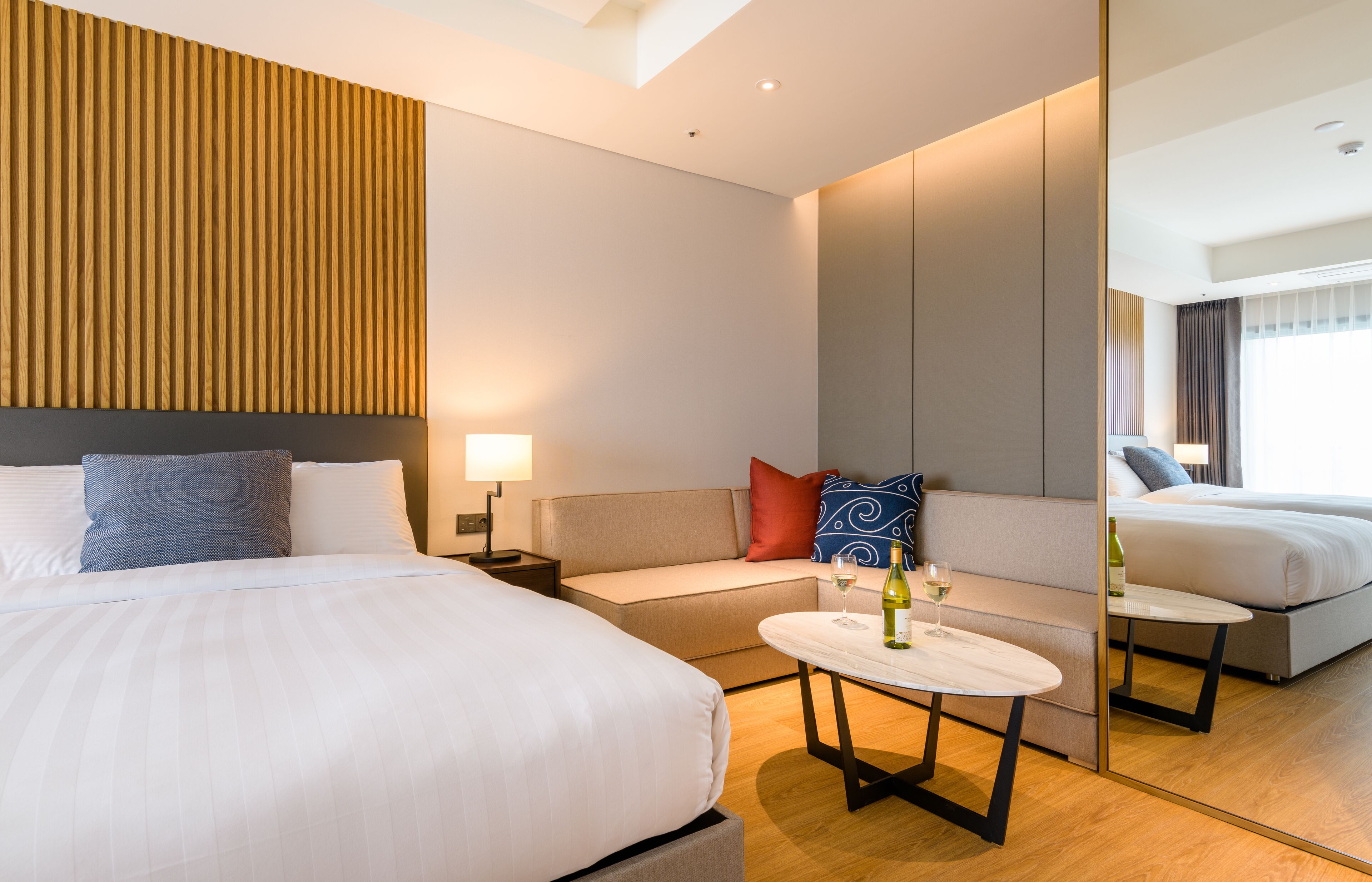 Una habitación de hotel moderna con una cama grande, un sofá, una mesita con una botella y un vaso, y un espejo que refleja la habitación.