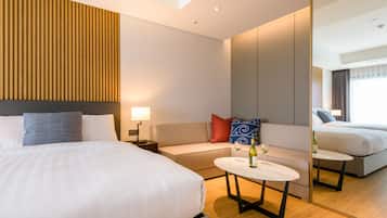 Una habitación de hotel moderna con una cama grande, un sofá, una mesita con una botella y un vaso, y un espejo que refleja la habitación.