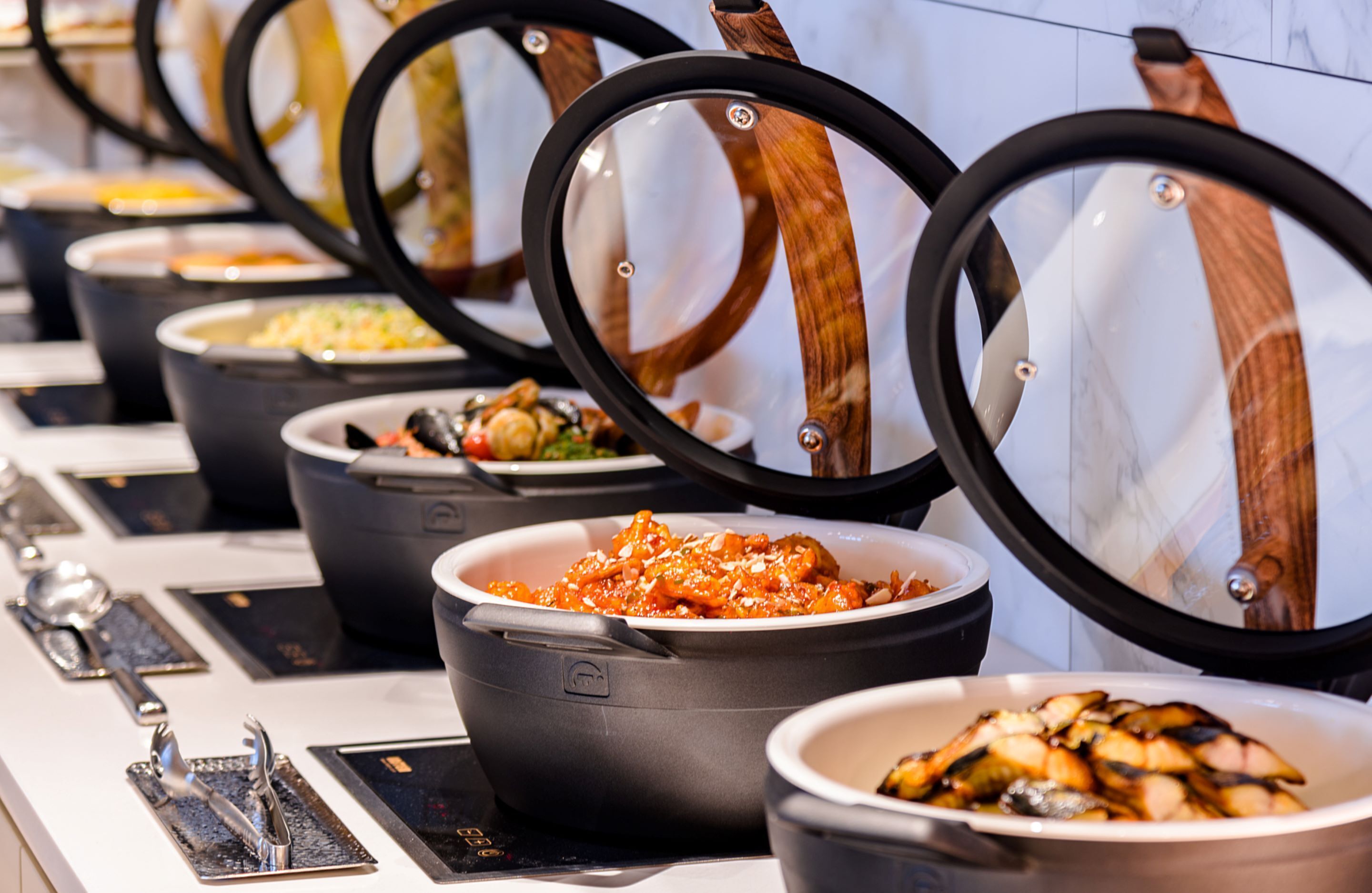 A buffet line with various dishes in black bowls, each with a wooden lid.