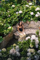 Una mujer sentada en un banco de piedra rodeada de exuberante follaje y flores de hortensia blancas.