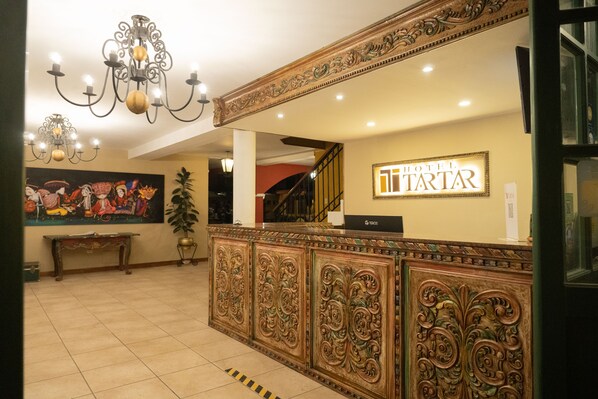 Zona de recepción de un hotel con paneles de madera decorativos, una araña, un cuadro y una escalera de fondo.