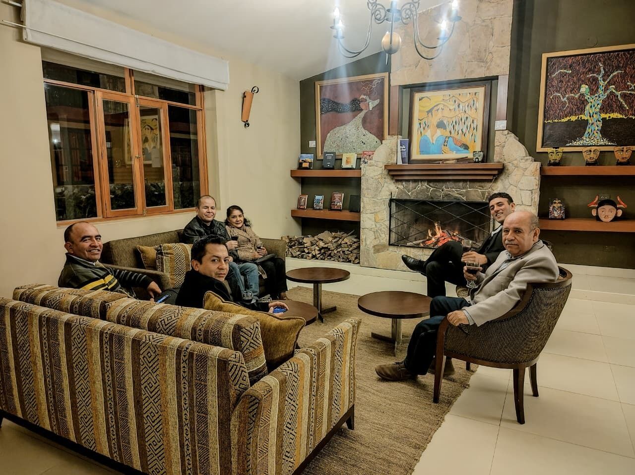 A cozy living room with a fireplace, patterned sofas, and a group of people seated around a coffee table.