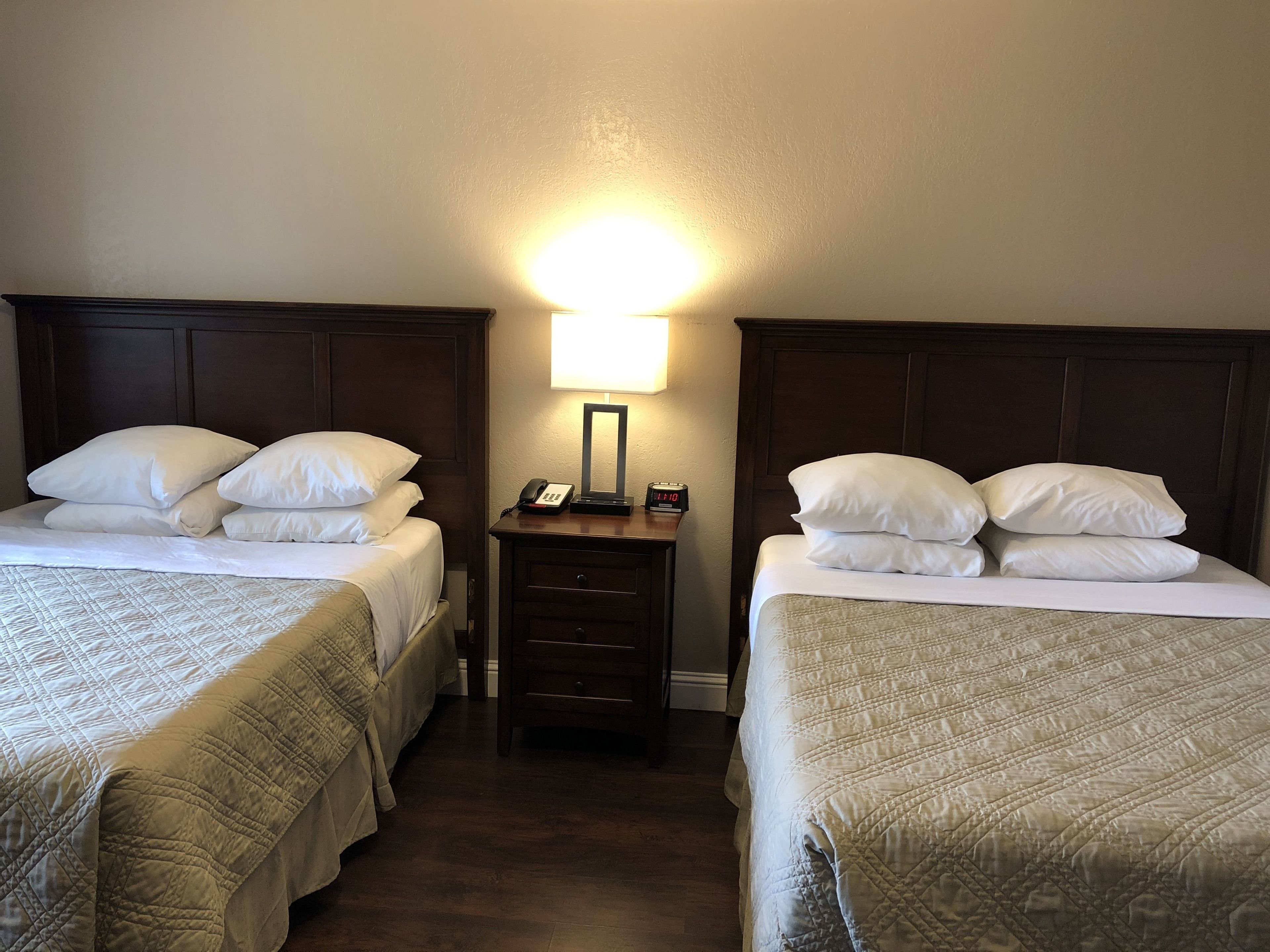 A hotel room with two beds, each with white linens and a quilted duvet. There is a nightstand between the beds with a lamp and a clock.
