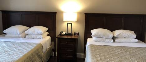 A hotel room with two beds, each with white linens and a quilted duvet. There is a nightstand between the beds with a lamp and a clock.