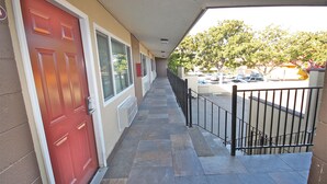 A covered walkway with a red door, a metal railing, and a parking lot visible in the background. - Budget Inn Lompoc (Lompoc)