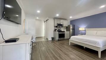 A compact hotel room with a bed, a desk, a kitchenette, and a TV mounted on the wall.
