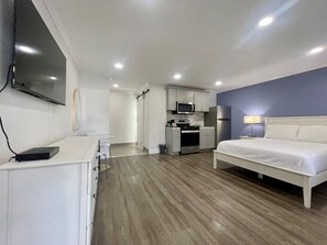 A compact hotel room with a bed, a desk, a kitchenette, and a TV mounted on the wall.