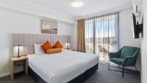 Habitación de hotel con una cama grande, dos mesitas de noche con lámparas, un sillón verde, un ventanal corredero a un balcón y un televisor de pared.