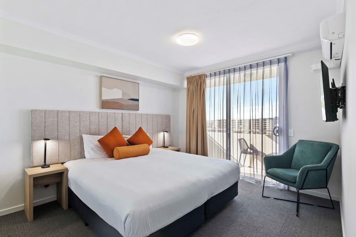 A hotel room with a large bed, two bedside tables with lamps, a green armchair, a sliding glass door to a balcony, and a mounted TV.