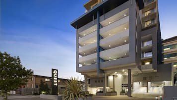 A multi-story hotel with a modern design, featuring a prominent "QUEST" sign, multiple balconies, and a well-lit entrance area.