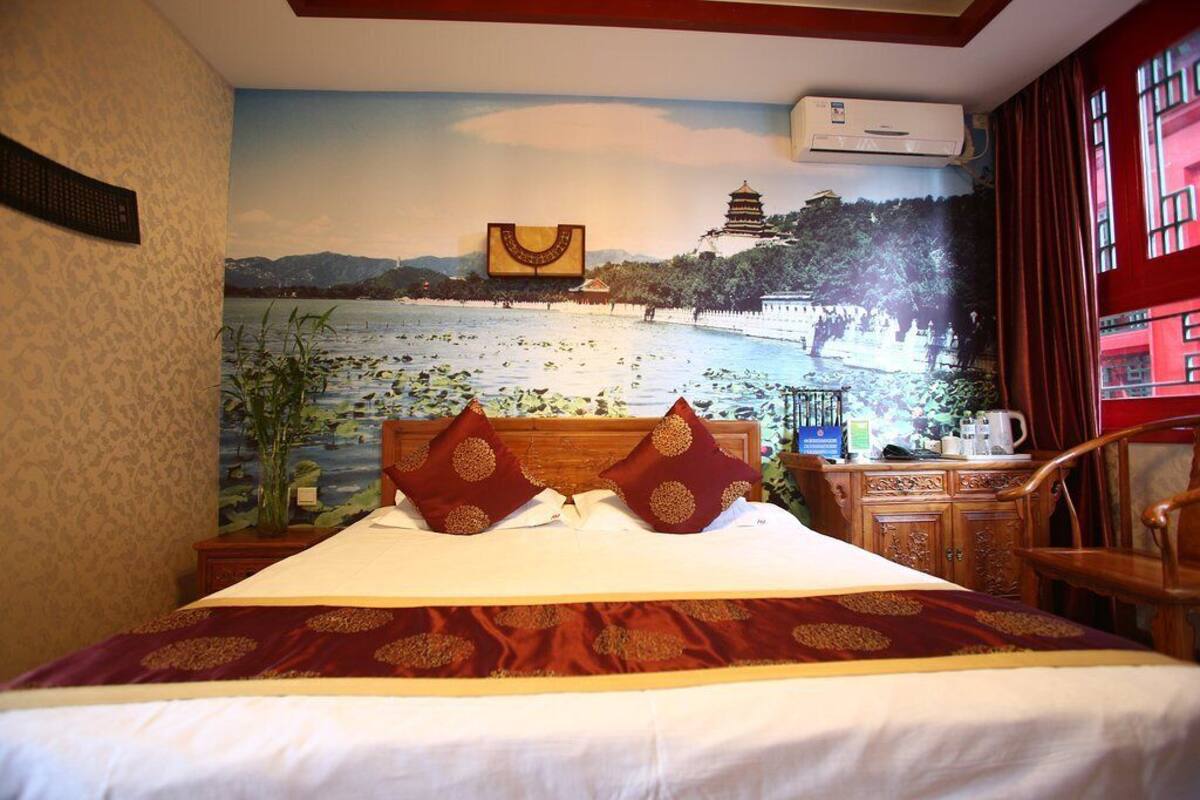 A hotel room with a bed, a nightstand, a wooden dresser, and a mural of a scenic landscape on the wall.