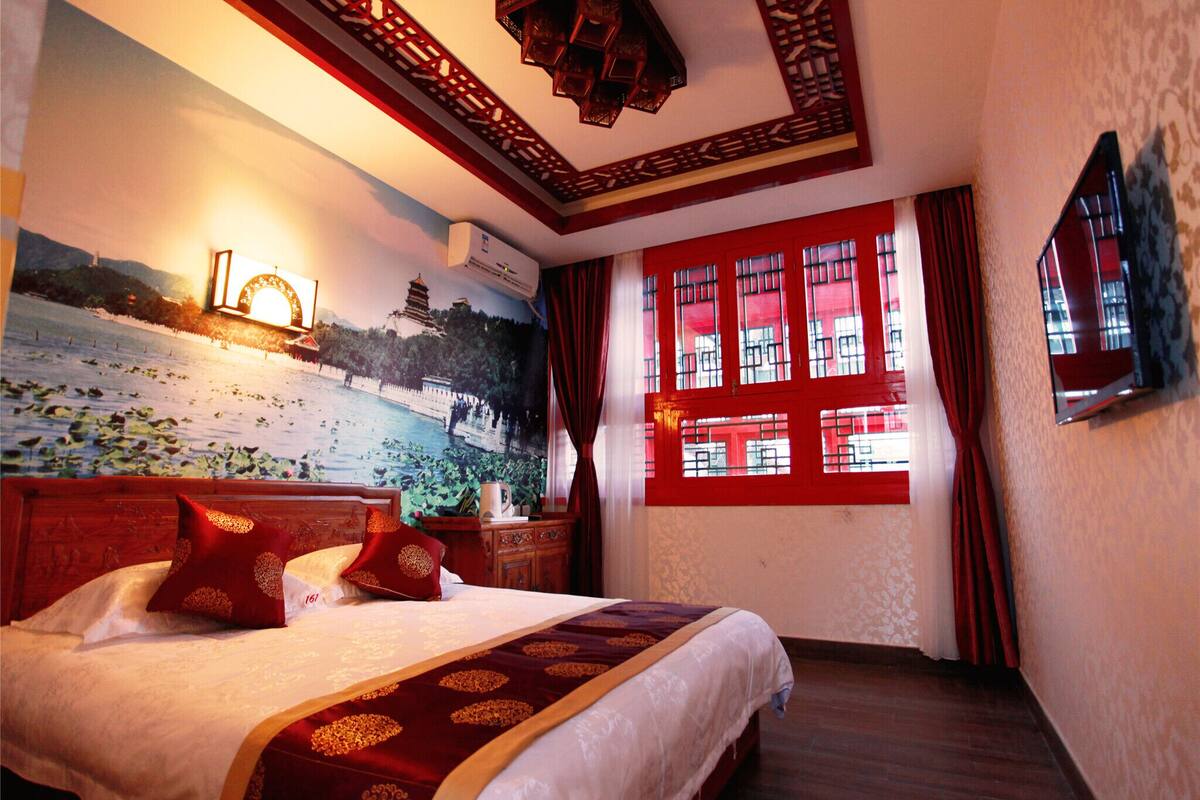 A hotel room with a large bed, a mural of a scenic landscape, a window with red curtains, and a traditional wooden ceiling design.